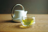 kleine transparente Glasteetasse neben weisser Teekanne auf Holzoberfläche - small transparent glass tea cup next to white teapot on wooden surface