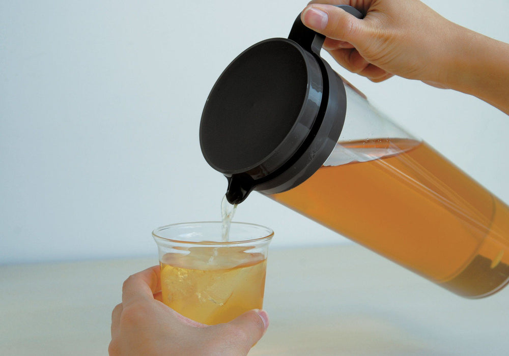 Hand gießt Eistee aus grosser transparenter Teekanne in kleine transparente Teetasse - hand pours ice tea from tall transparent tea jug into small transparent teacup